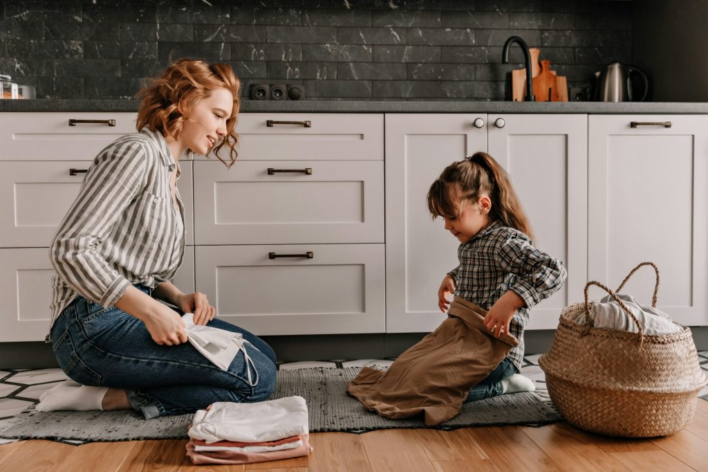 Young mother and her cute daughter gently fold clothes sitting on kitchen floor